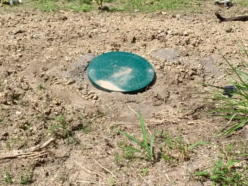 Couvercle vert d'une cuve enterrée dans un terrain de terre sèche, maison rose en arrière-plan