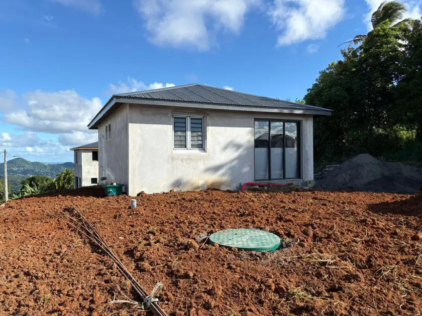 Maison neuve aux murs bruts, couvercle vert d'une cuve enterrée dans la terre rouge devant la baie vitrée