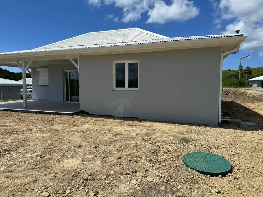 Maison neuve au crépi gris, couvercle vert d'une cuve enterrée dans le terrain nu