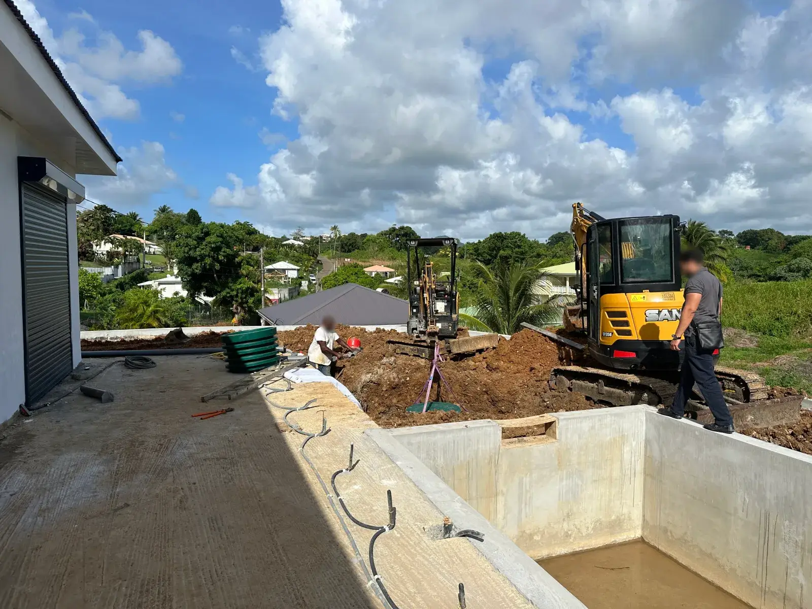 Deux mini-pelles et la fouille à côté d'une piscine en construction, la cuve suspendue au-dessus du trou