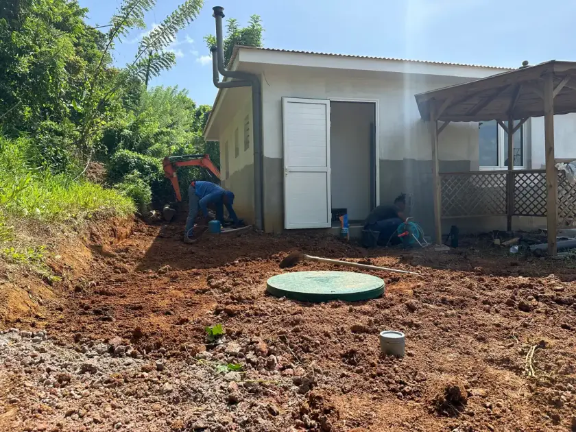Deux ouvriers au travail devant une maison, couvercle vert de cuve au centre du terrain remué, mini-pelle en arrière-plan