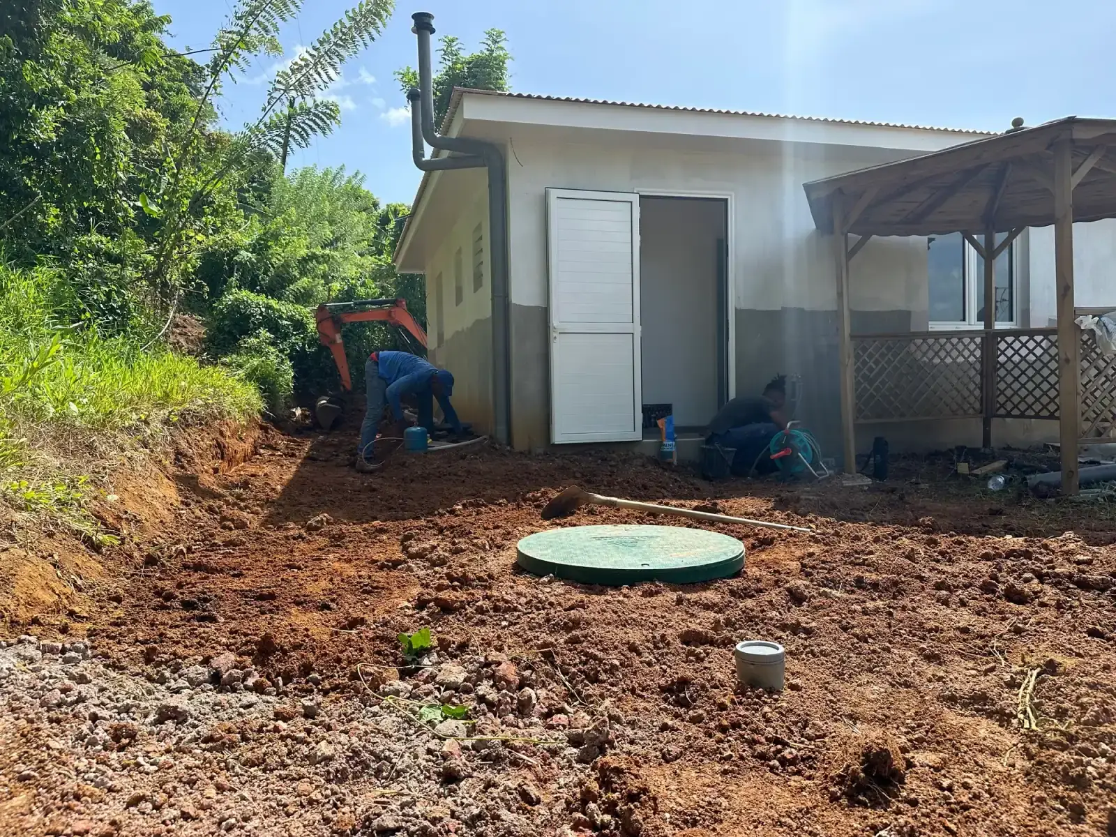 Deux ouvriers au travail devant une maison, couvercle vert de cuve au centre du terrain remué, mini-pelle en arrière-plan