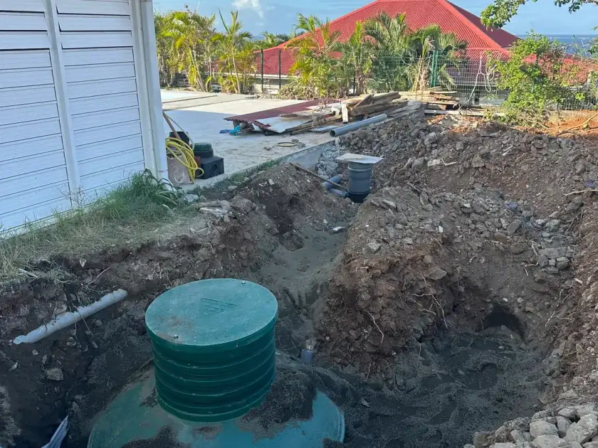 Cuve verte en cours de pose dans une fouille, avec ses rehausses, à côté d'une maison créole, mer à l'horizon