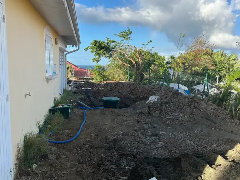 Terrain remué le long d'une maison jaune, couvercle vert de la cuve posée et gaine bleue, mer au loin